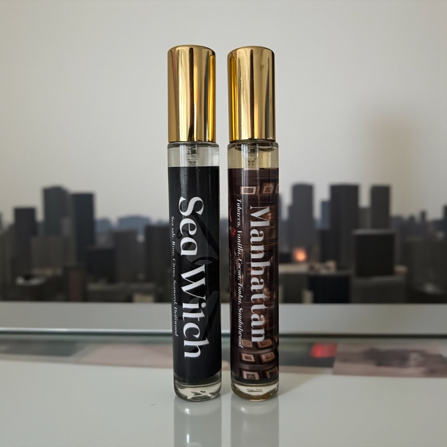 Two perfume bottles labeled 'Sea Witch' and 'Manhattan' on a reflective surface with a cityscape background.