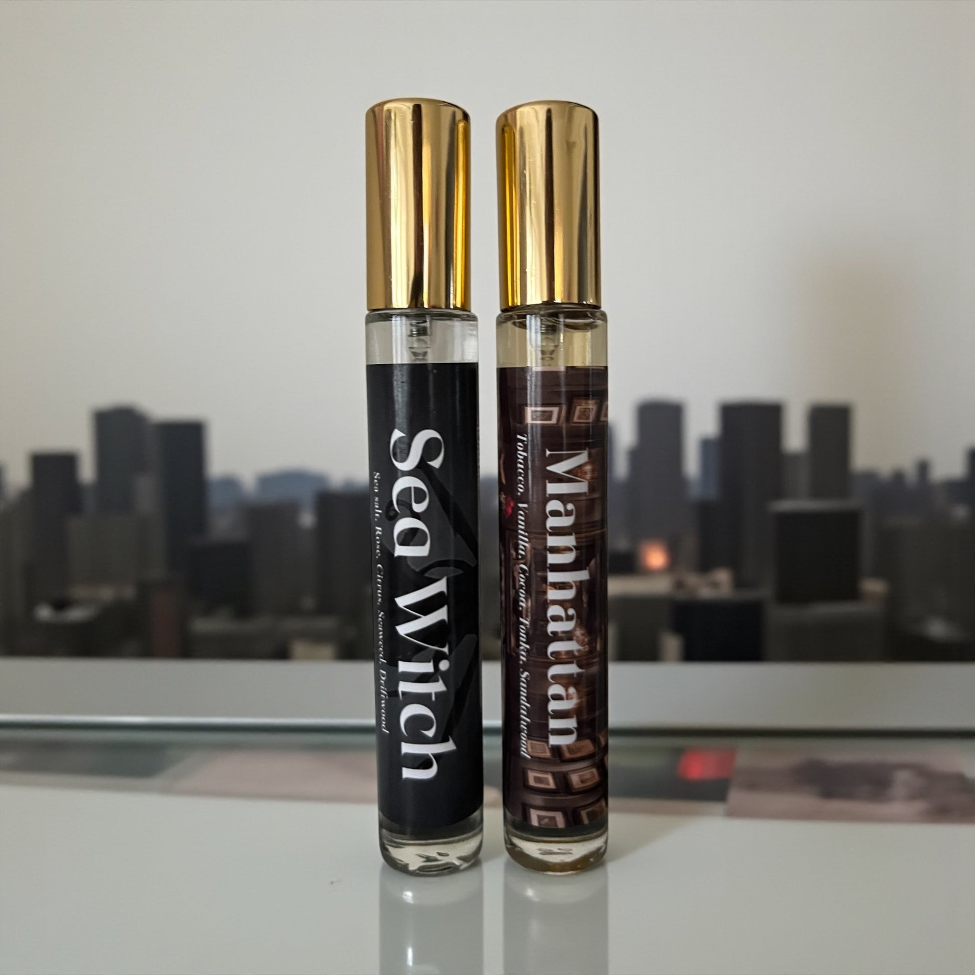 Two perfume bottles labeled 'Sea Witch' and 'Manhattan' on a reflective surface with a cityscape background.