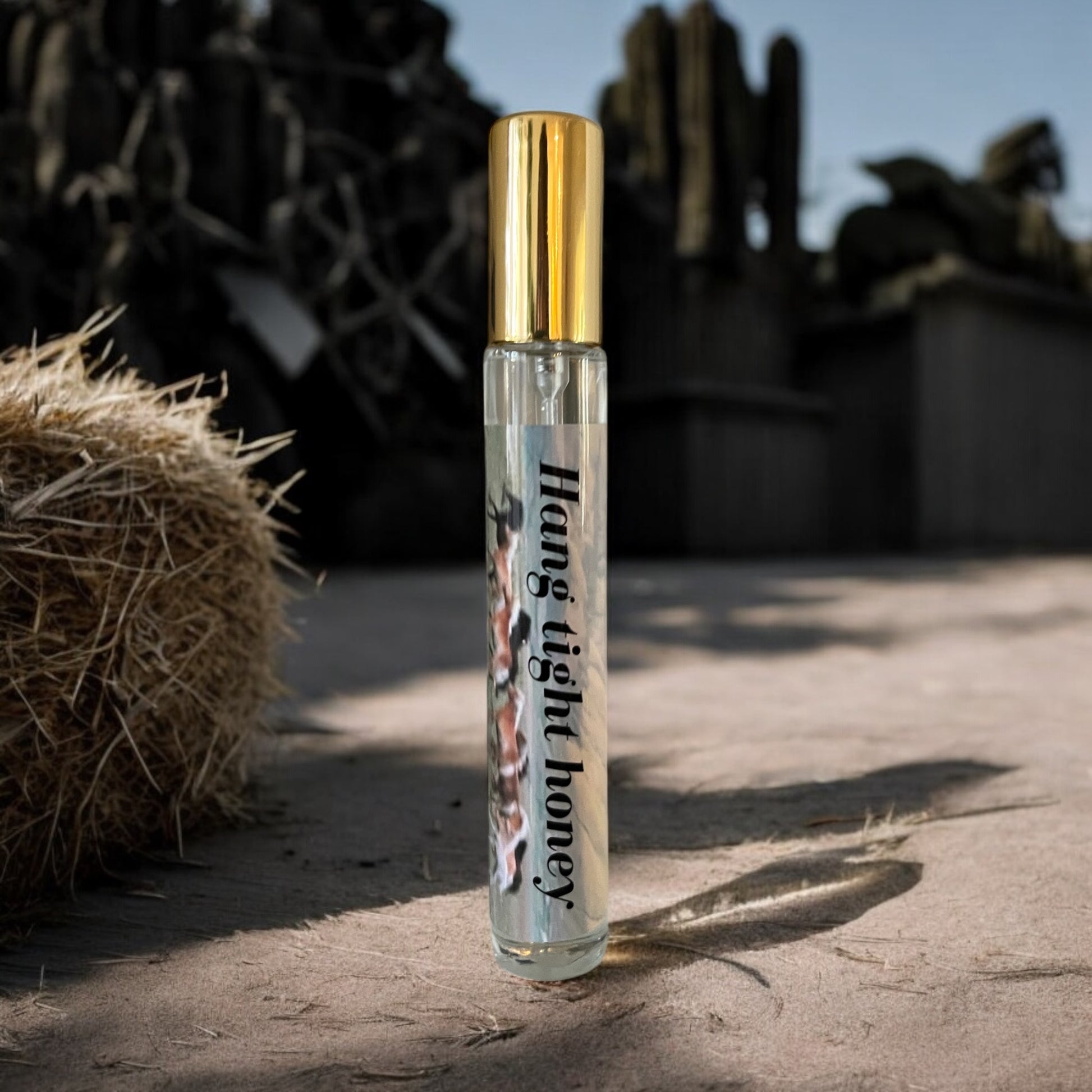 A bottle of Hang tight honey perfume on the ground with hay and cactus in the background evoking the grassy fresh scent