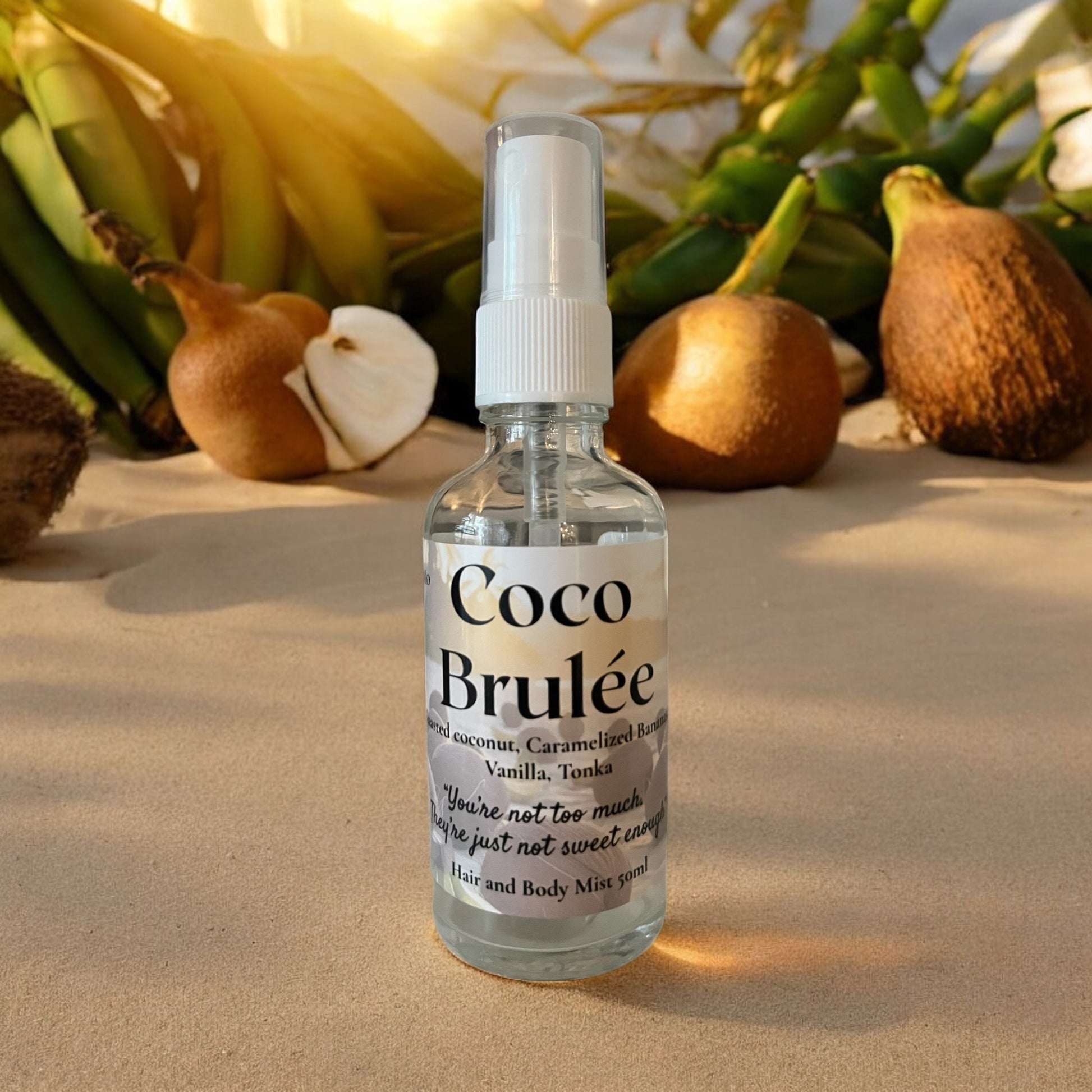 Coco Brulee tropical gourmand  hair and body mist bottle sitting in the sand with coconuts and green stems