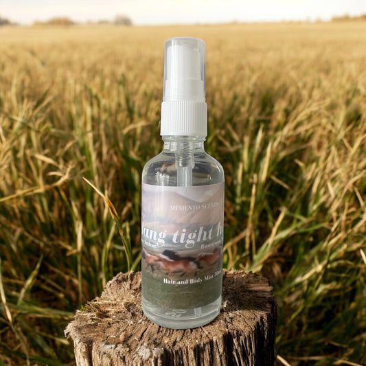 A bottle of Hang tight honey hair and body mist on a tree stump in a field of hay evoking the grassy earthy fresh scent