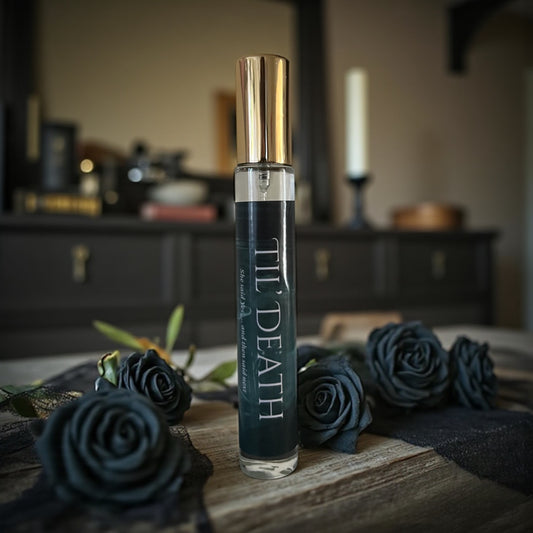 Perfume bottle labeled 'Til Death' surrounded by black roses on a wooden surface.