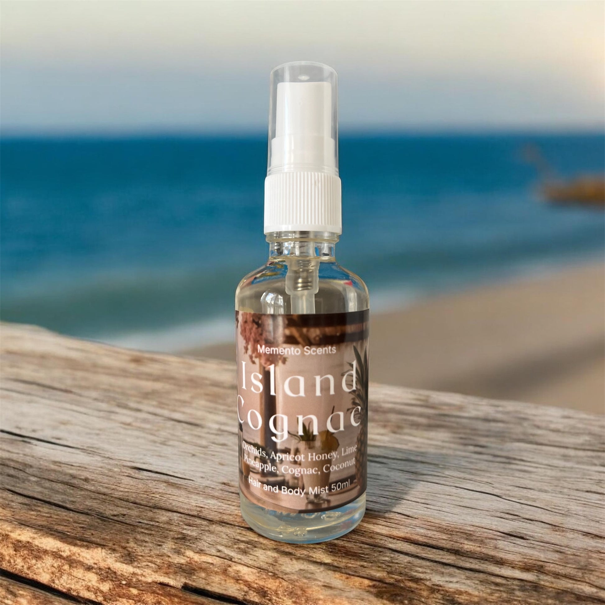 A glass spray bottle of hair and body mist on a wood ledge with the ocean behind named Island Cognac